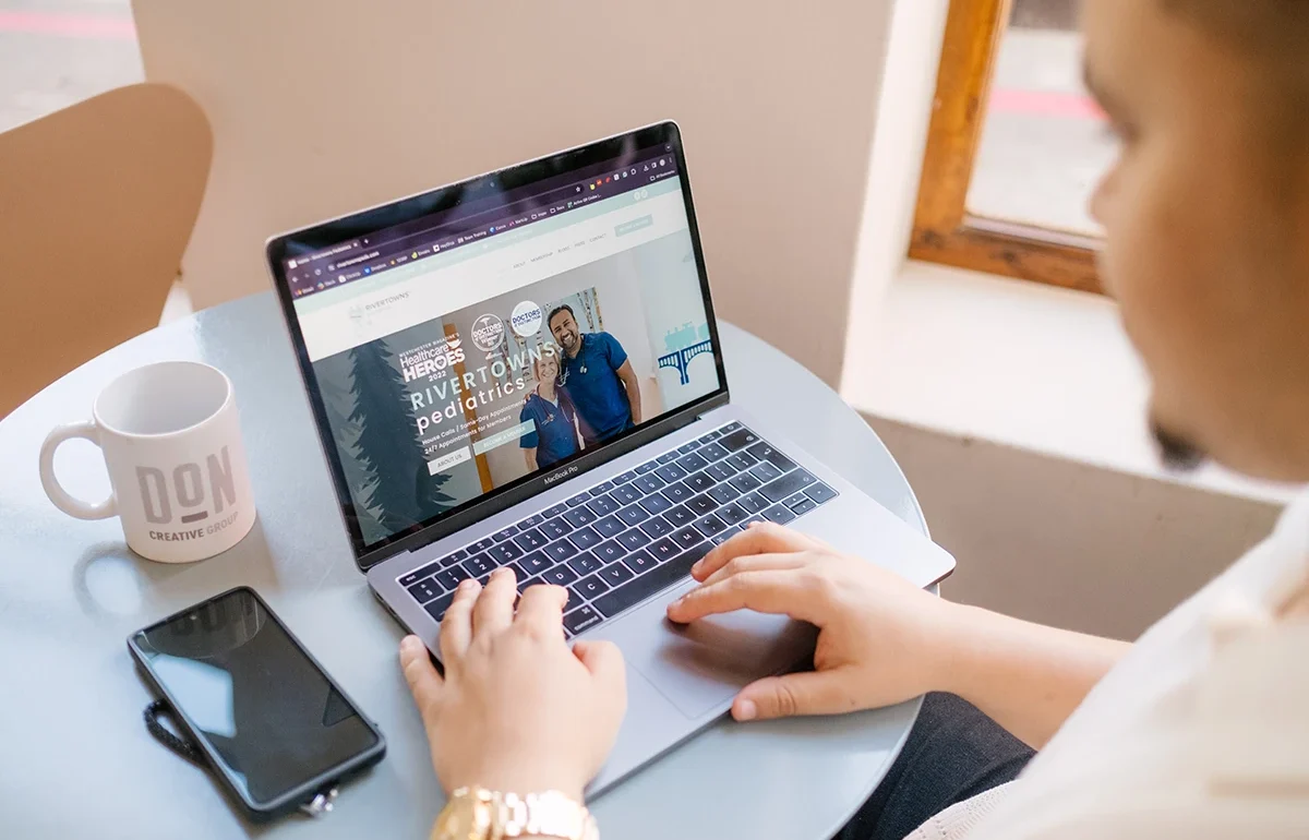 Person browsing the Rivertowns Pediatrics website on a laptop at a round table, with a Don Creative Group mug and phone nearby.