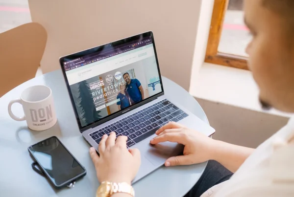 Person browsing the Rivertowns Pediatrics website on a laptop at a round table, with a Don Creative Group mug and phone nearby.