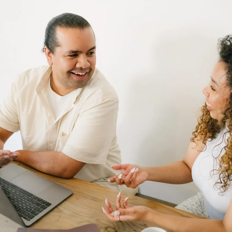 Two coworkers smiling and having a lively conversation at a desk with a laptop during a collaborative work session.