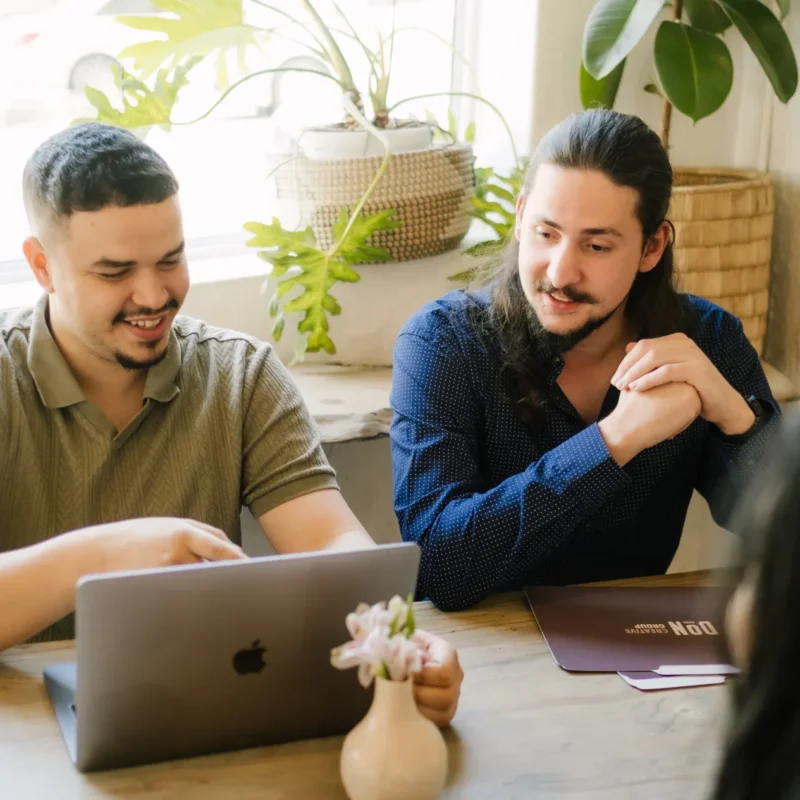 Website developer, Douglas and Jnr Social Media Manager, Jacques, sitting accross from two unidenifiable people on a desk while looking at designs on a macbook