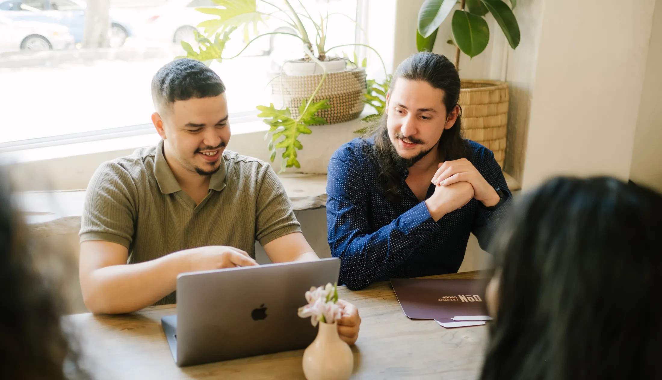Website developer, Douglas and Jnr Social Media Manager, Jacques, sitting accross from two unidenifiable people on a desk while looking at designs on a macbook