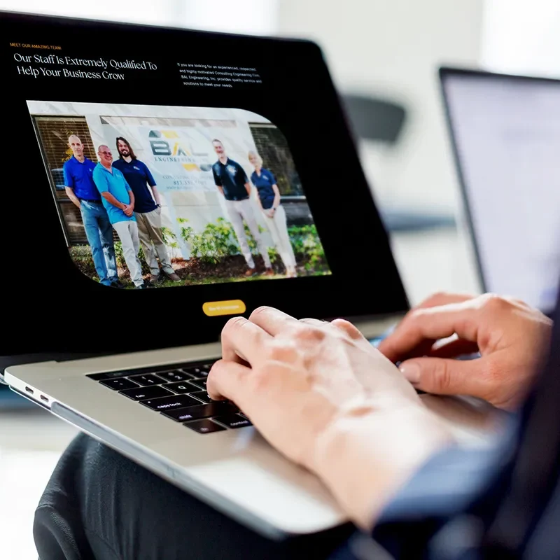 Person viewing a business website on a laptop, showcasing a team photo with the headline “Our Staff Is Extremely Qualified To Help Your Business Grow.
