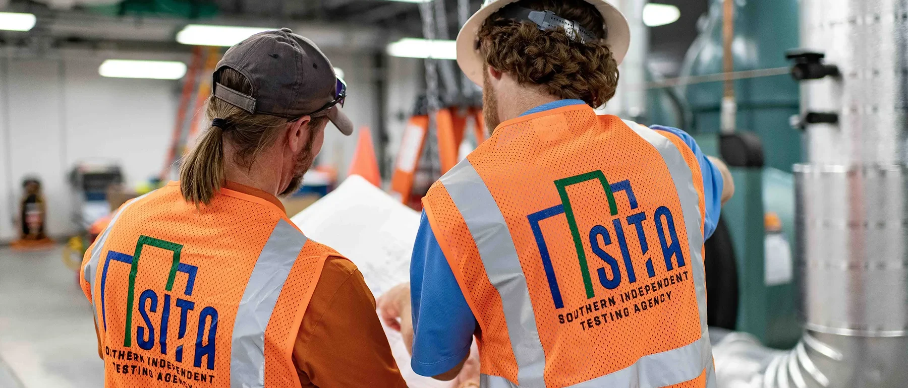 Two construction professionals wearing safety vests review plans inside a mechanical room during HVAC testing and inspection