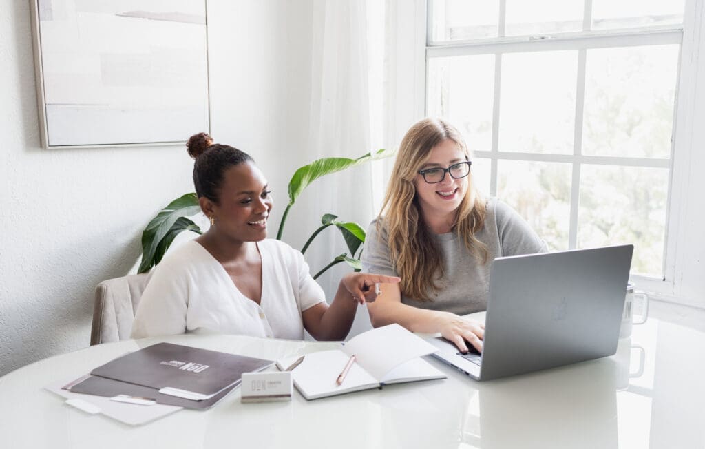 Two women collaborating at a desk with a laptop, notebook, and Don Creative Group materials.