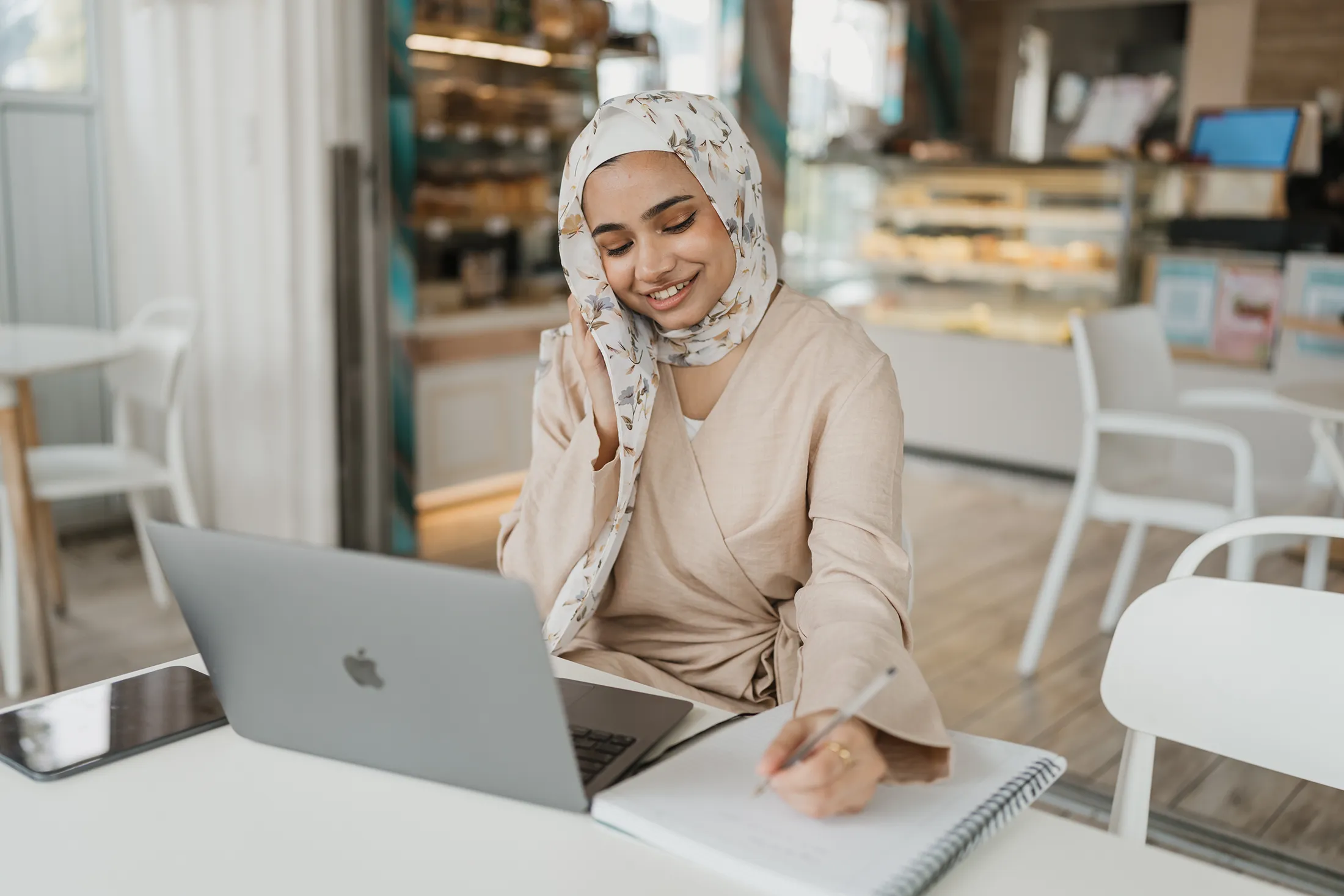 Woman working on a laptop while writing notes at a cafe, representing social media planning, content strategy development, and digital marketing workflow
