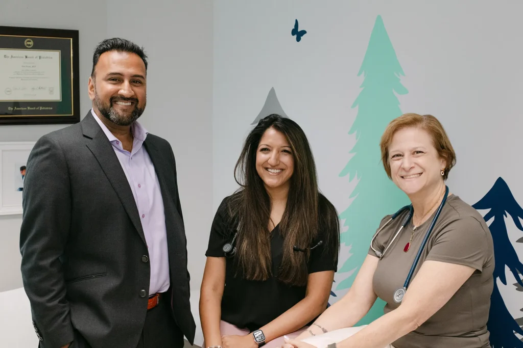 Pediatric Care Team Smiling in Clinic Environment A pediatric care team of three professionals smiling together in a child friendly exam room, standing in front of colorful wall art during a clinic visit.