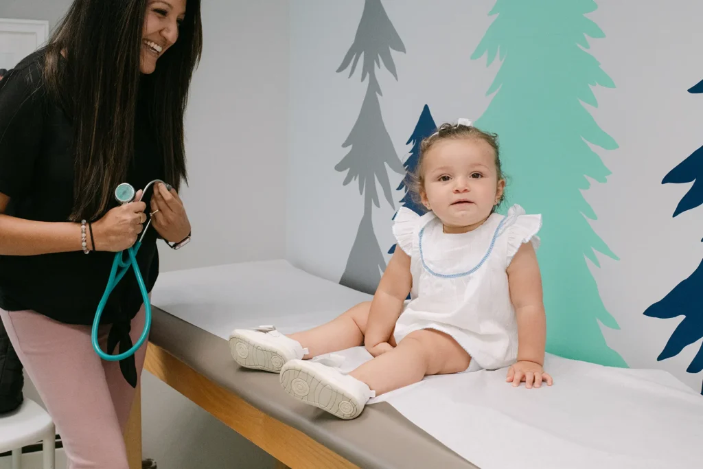 Toddler Sitting on Exam Table During Pediatric Checkup A toddler sits on an exam table while a smiling pediatric provider holds a stethoscope in a bright, child friendly exam room with colorful wall art.