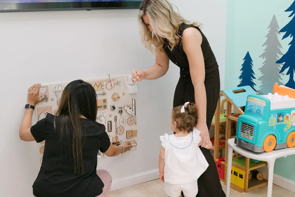 Toddler Playing with Sensory Wall in Pediatric Office A toddler interacts with a sensory activity wall while a mother and pediatric staff member assist in a brightly decorated pediatric playroom with toys and colorful wall art.