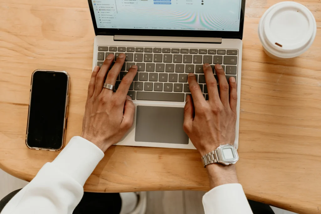 Hands typing on a laptop keyboard at a wooden desk with phone and coffee nearby