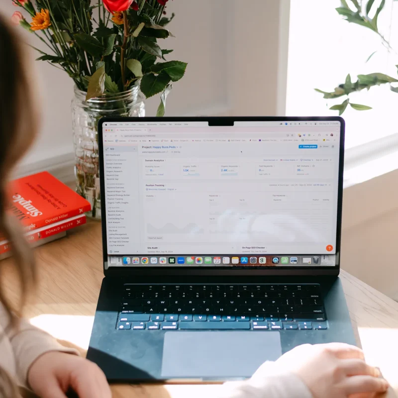 Person reviewing SEO analytics and keyword performance dashboard on a laptop at a home office desk with flowers and books.