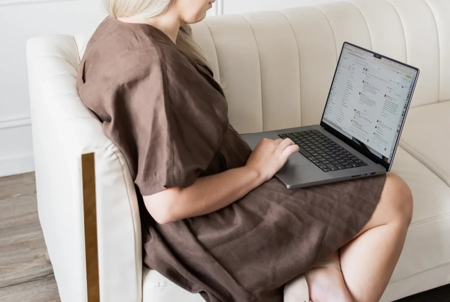 Professional woman seated on a couch working on a laptop, reviewing emails and digital communications in a bright, modern office setting.