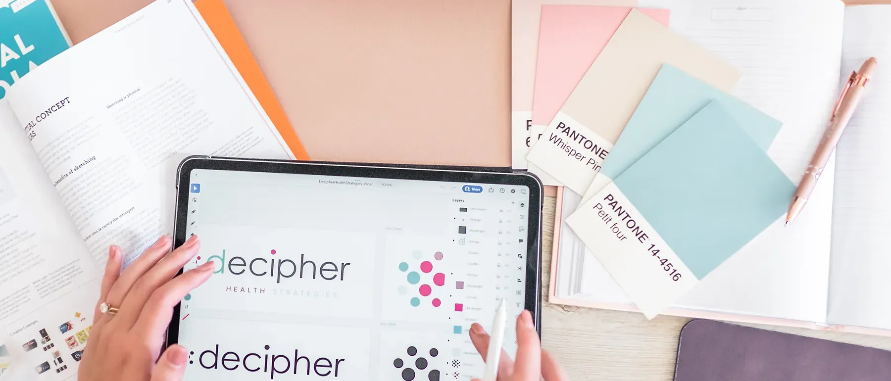 xDesigner reviewing Decipher Health logo concepts on tablet beside color swatches and notebooks