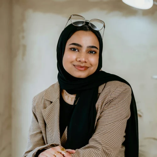 Team member wearing black hijab and plaid blazer, smiling indoors at office