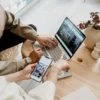 Two professionals reviewing digital content, one using a laptop and the other holding a smartphone, collaborating at a wooden table with minimalist decor.