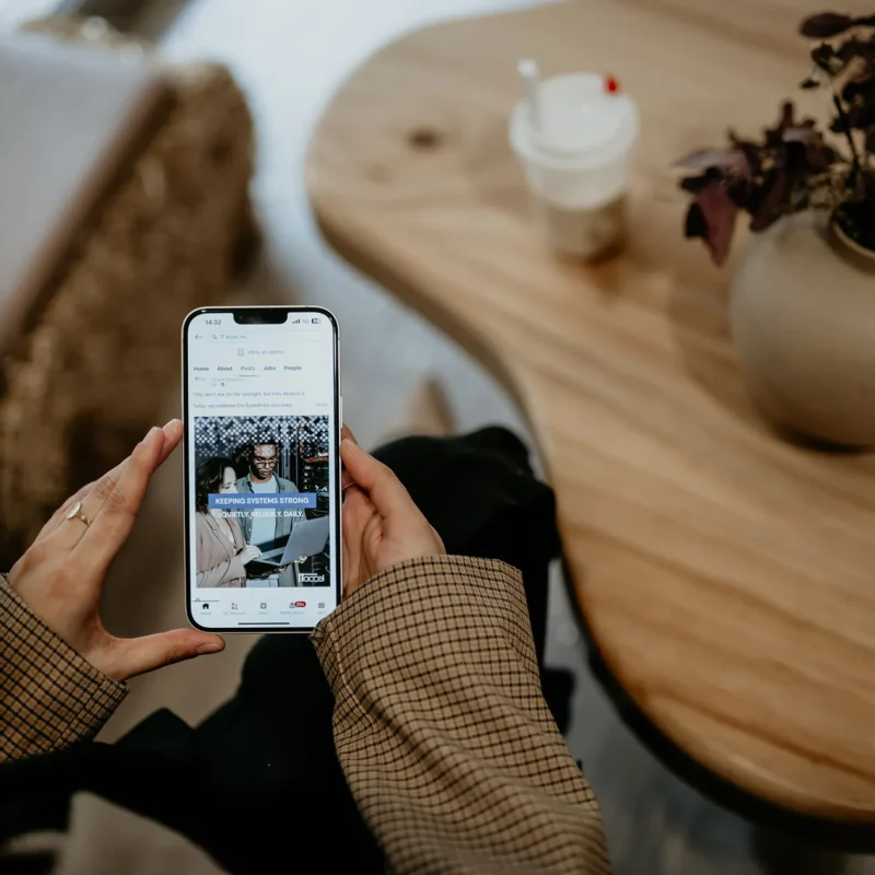 Person holding a smartphone viewing a business social media post while seated at a wooden table with a coffee cup and decorative vase nearby.