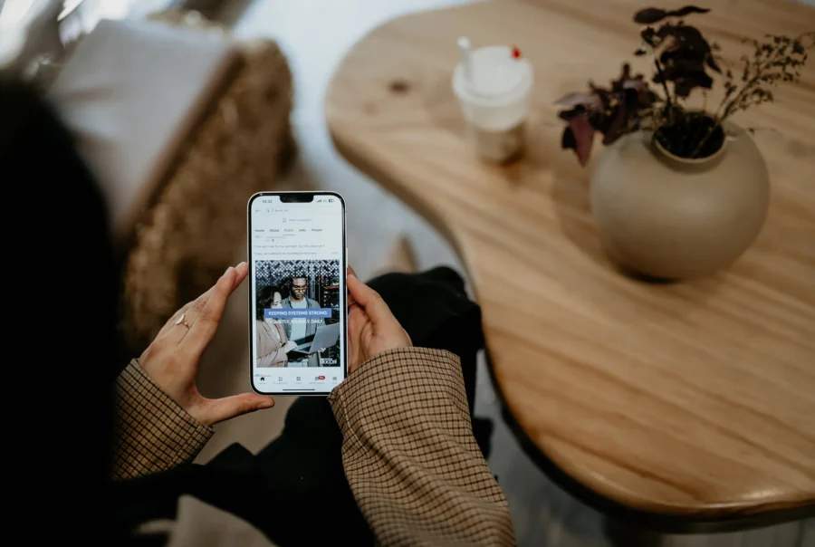 Person holding a smartphone viewing a business social media post while seated at a wooden table with a coffee cup and decorative vase nearby.