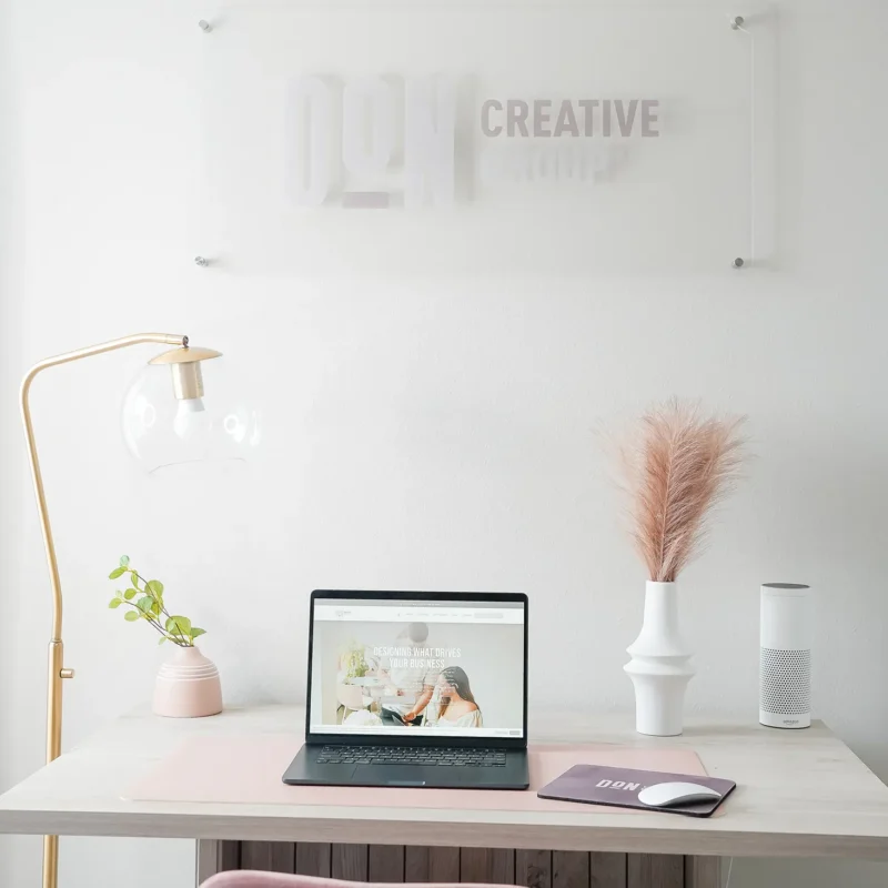Minimalist office workspace with a laptop on a desk, soft pink chair, desk lamp, decorative plants, and a Don Creative Group wall sign in the background.