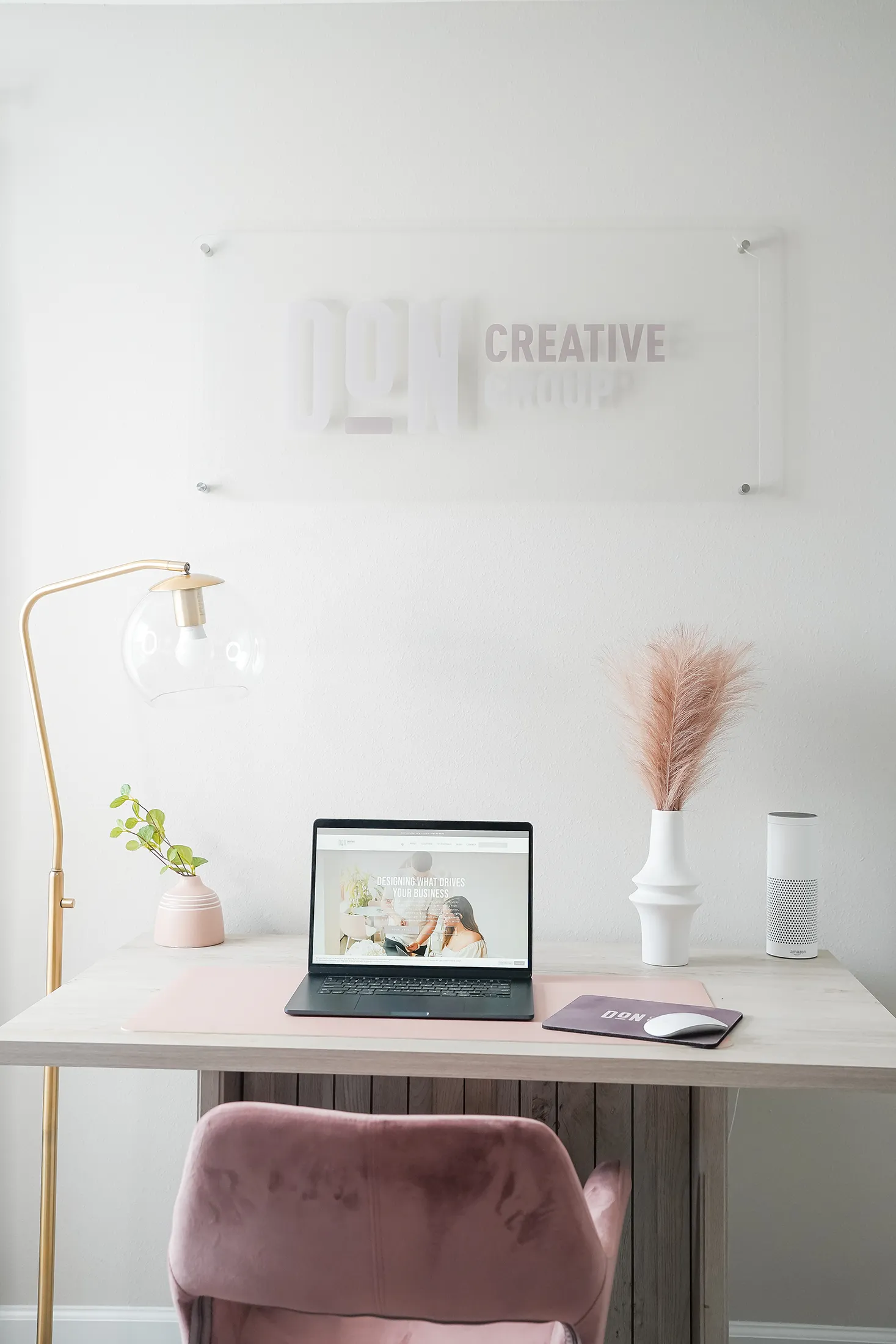 Minimalist office workspace with a laptop on a desk, soft pink chair, desk lamp, decorative plants, and a Don Creative Group wall sign in the background.
