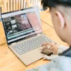 Person editing a video project on a laptop at a wooden table, with a video timeline, media assets, and editing tools visible on the screen.