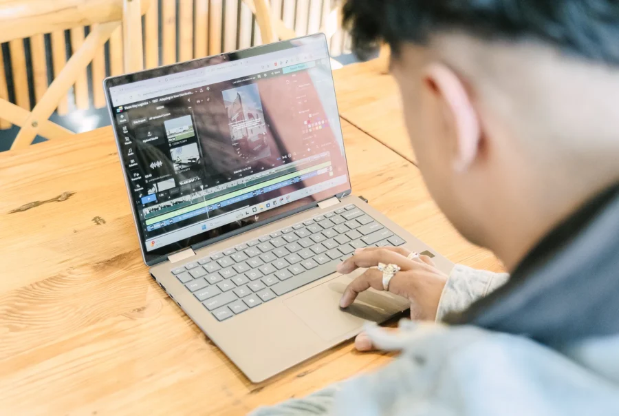 Person editing a video project on a laptop at a wooden table, with a video timeline, media assets, and editing tools visible on the screen.