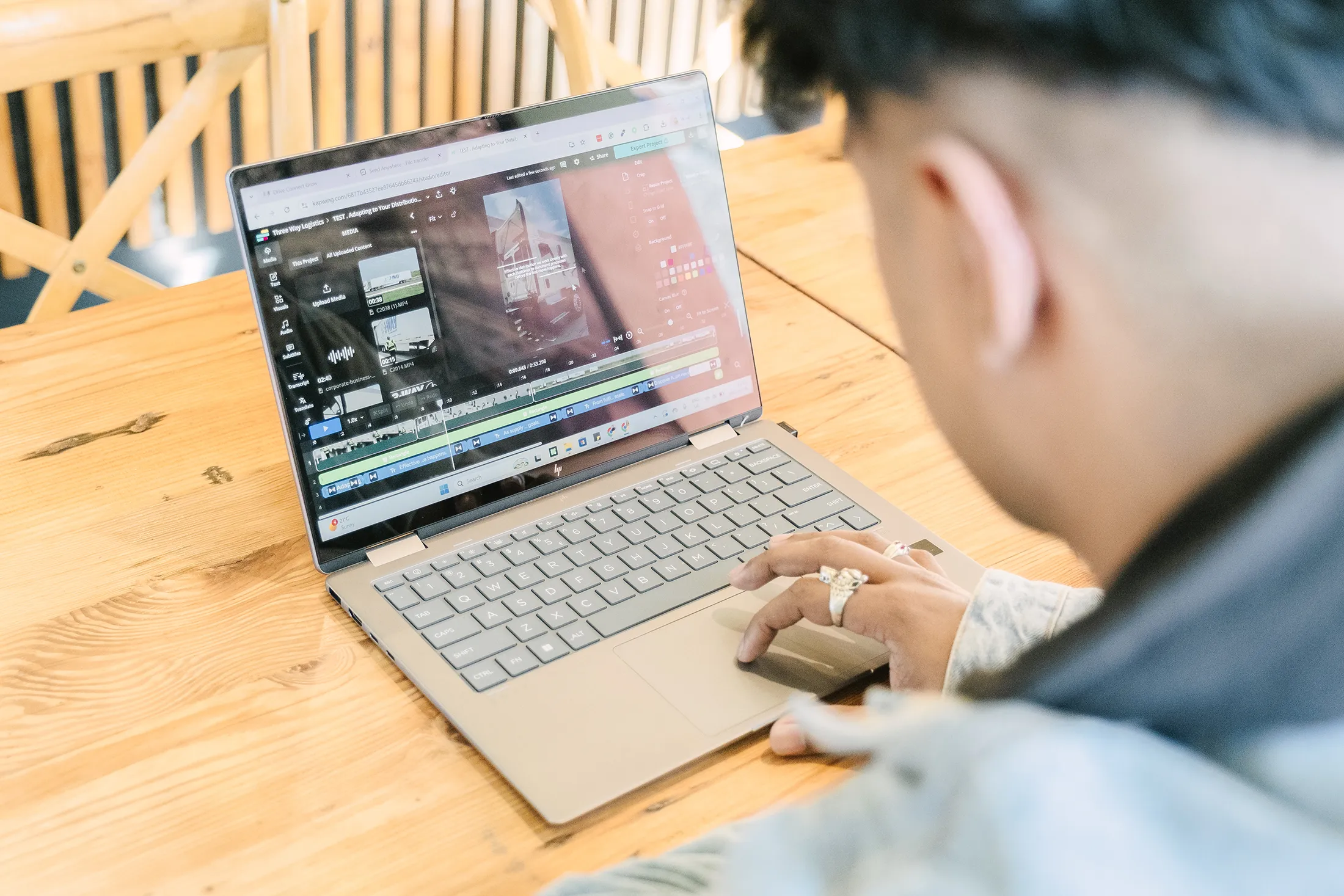Person editing a video project on a laptop at a wooden table, with a video timeline, media assets, and editing tools visible on the screen.