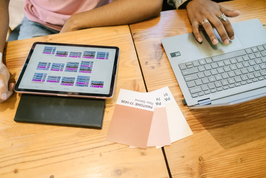 Two people working at a wooden table with a tablet showing a content schedule, a laptop, and color swatch cards laid out for branding and design planning.