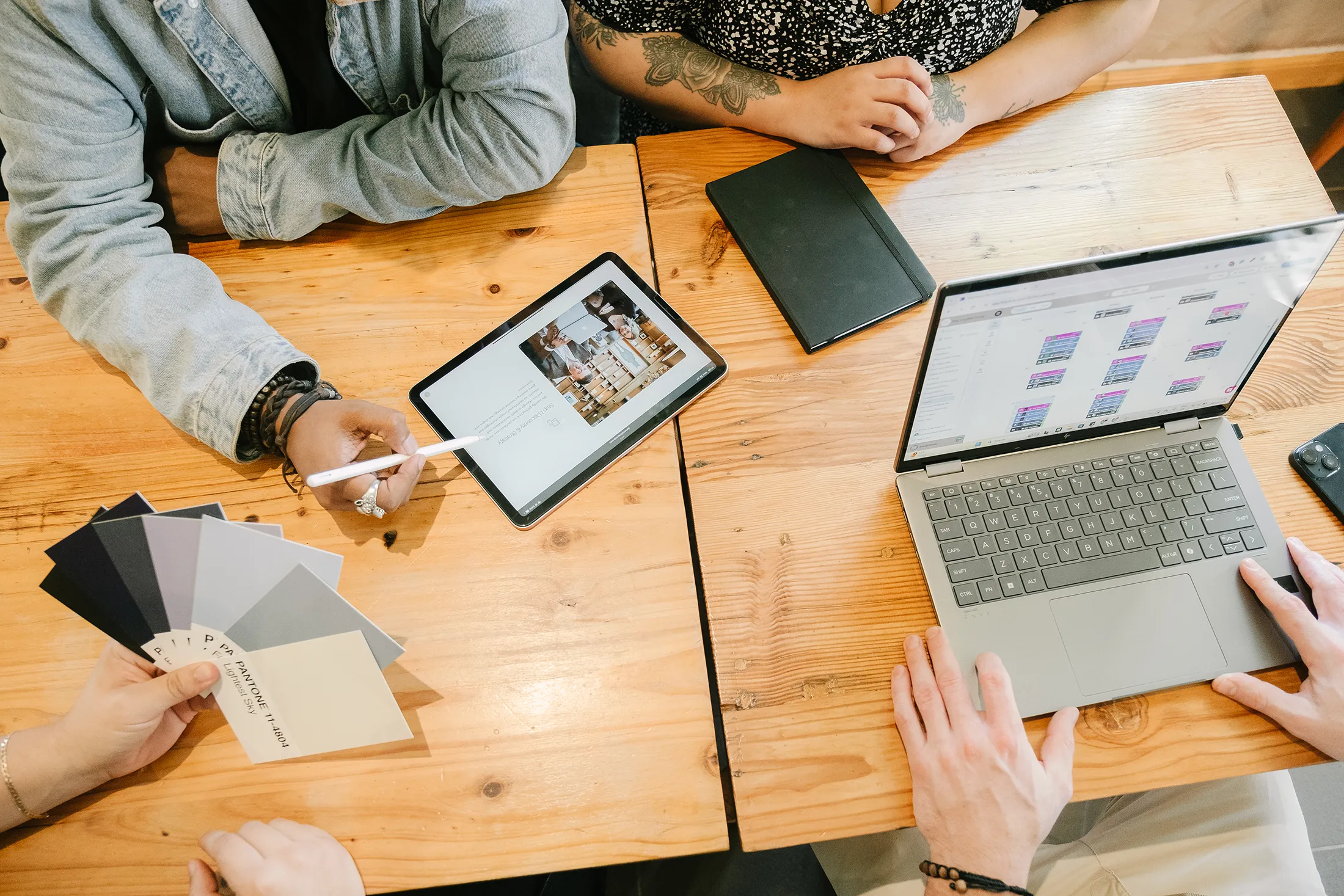 Team collaborating at a wooden table with a tablet displaying a website layout, a laptop showing content planning, and Pantone color swatches used for brand identity and rebranding work.